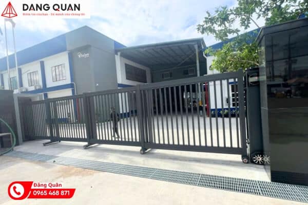 Cổng xếp phân đoạn thân nhôm màu xám đậm, các đoạn khung song đứng nối khớp, đặt trước nhà xưởng