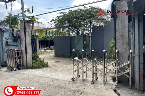 Cổng xếp inox kéo tay đang kéo dở ngang lối vào nền bê tông, hai bên là trụ cổng