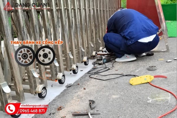 Thợ ngồi thay bánh xe dưới thân cổng xếp inox, chữ thay bánh xe cổng xếp và hai bánh xe mẫu in trên ảnh