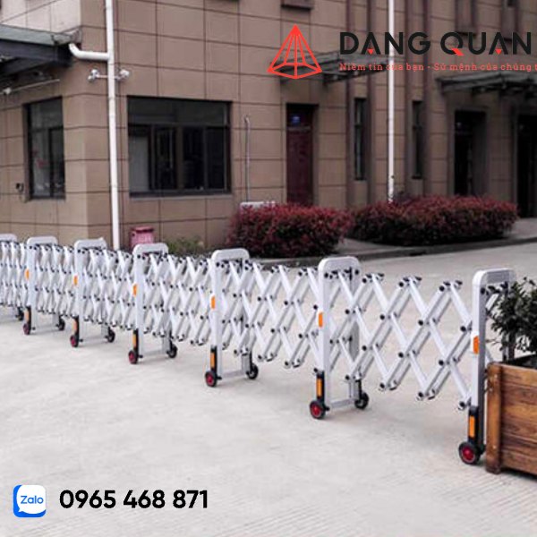 Cổng xếp nhôm kéo tay màu trắng bạc kéo dài dọc lối vào trước một tòa nhà, chân trụ có bánh xe